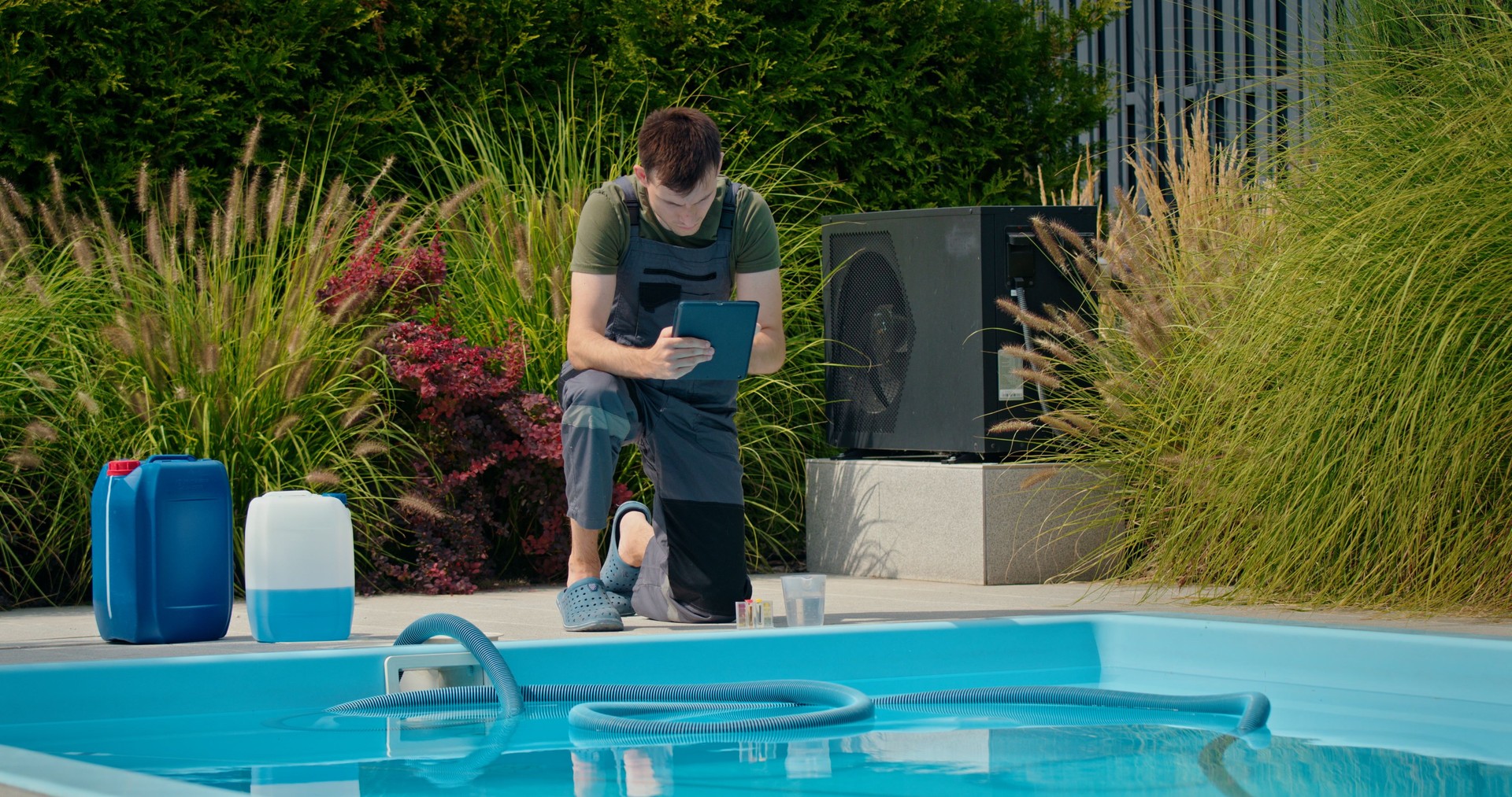 Pool technician checking data on a tablet beside a swimming pool with chemical containers and lush greenery.